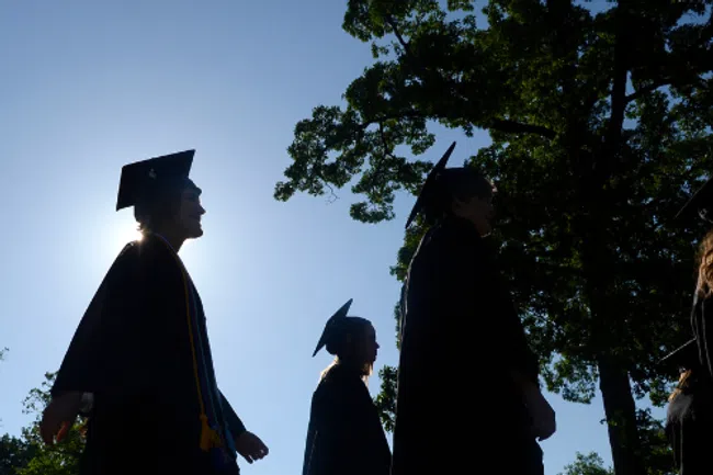 Silhouetted graduates in caps and gowns walking outdoors with trees and clear sky in background at graduation ceremony.