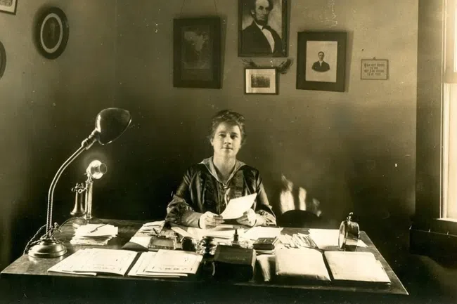 Early 20th-century woman sitting at desk with papers and vintage telephone under portraits on wall