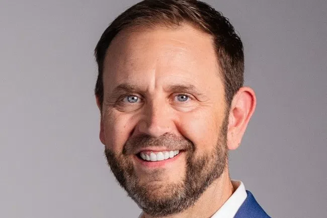 Smiling man with beard wearing a blue blazer and white shirt against a plain gray background