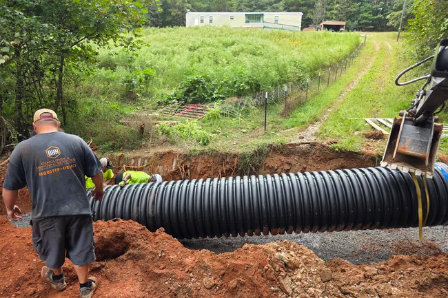 Workers installing large black corrugated drainage pipe in a rural area near a mobile home and dirt road.