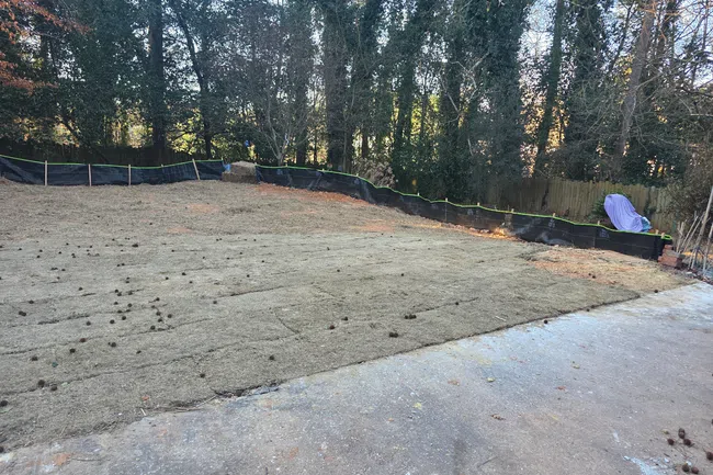 Newly laid sod covering an open yard area bordered by a tree line and black erosion control fencing.