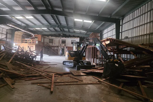 Indoor construction site with excavator clearing wooden debris inside a large warehouse with workers present
