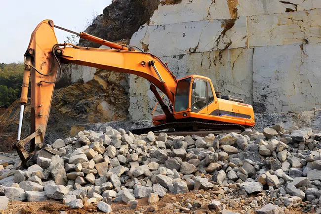 Orange excavator working on rocky terrain in a quarry with steep rock walls and scattered stones.