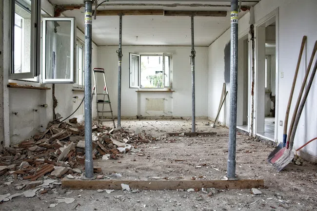 Room under renovation with open windows, scattered debris, metal support pillars, and construction tools visible.