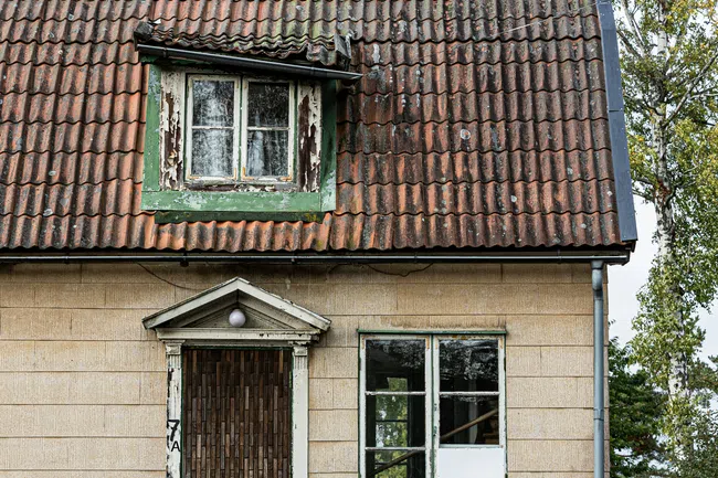 Old beige house with weathered red tile roof, peeling paint, a broken window, and a pink scooter outside.