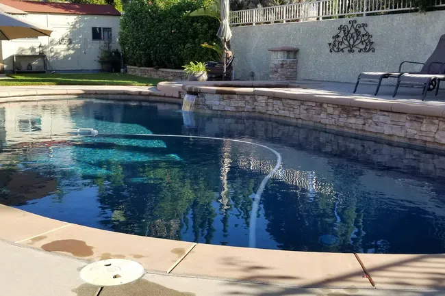 Clear blue backyard swimming pool with stone wall, lounge chairs, and large umbrella on sunny day.