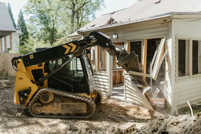 Yellow skid steer loader demolishing a house wall on a residential construction site with dirt and debris.