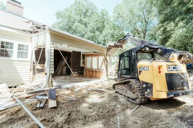 Yellow JCB excavator demolishing a house with debris and exposed interior under bright daylight.