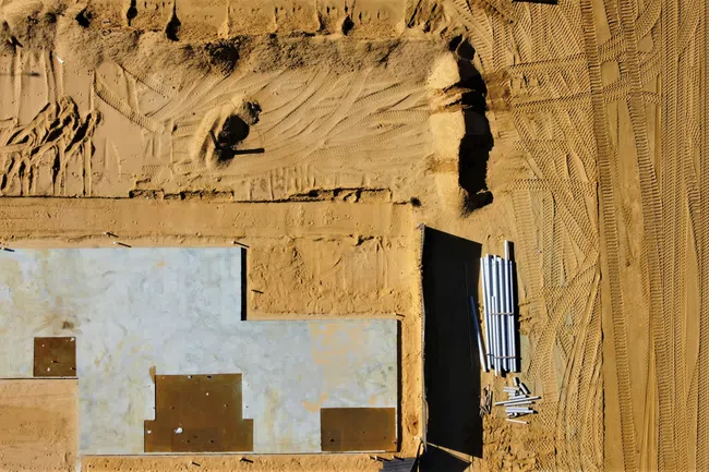 Aerial view of a construction site with foundation slab, dirt, and construction pipes on sandy ground.