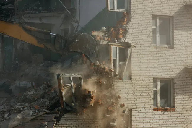 Excavator demolishing a brick building wall with windows during daytime, debris and dust flying.