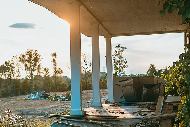 Broken wooden porch with white pillars in sunlight, surrounded by grass and overgrown plants in rural area.
