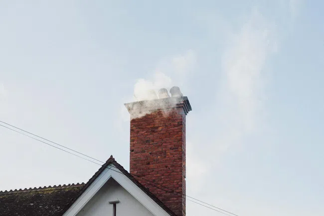 Brick chimney on a house with smoke rising against a clear blue sky during daytime.