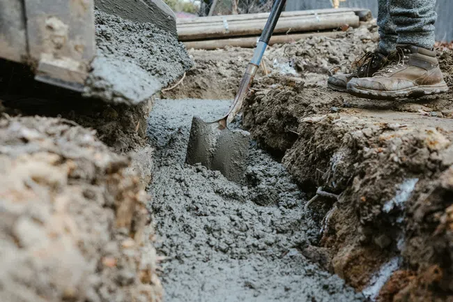 Worker pouring and leveling wet concrete into a trench with a shovel at a construction site outdoors.