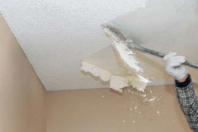 Hand removing popcorn ceiling texture with scraper tool in an interior beige room renovation.