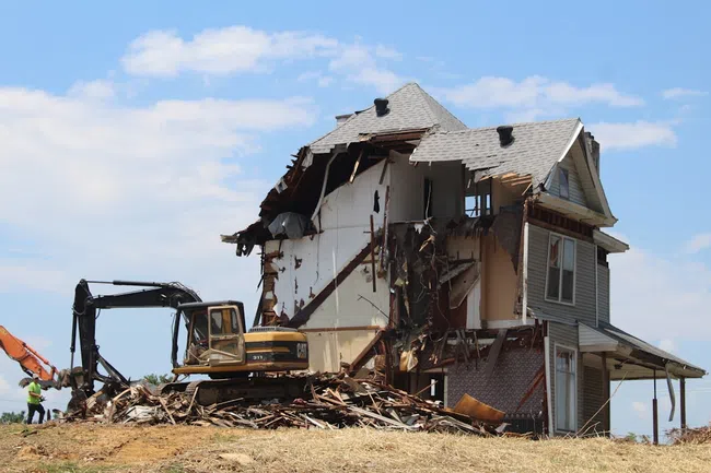 Partially demolished two-story house with exposed interior walls and construction machinery on site under blue sky.