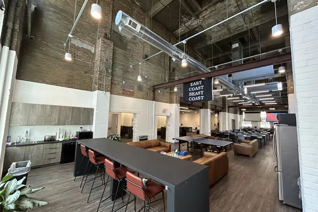 Spacious modern office lounge with high ceilings, industrial ductwork, kitchen area, bar seating, and ping pong tables.