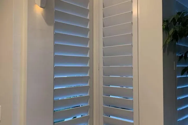White plantation shutters cover corner windows in a modern, well-lit interior space with minimal decor.