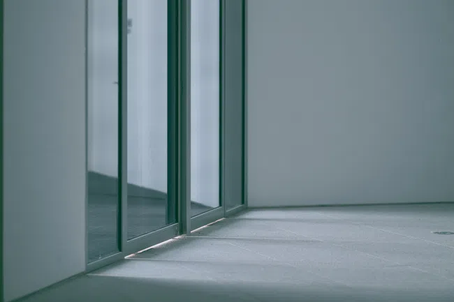 Empty modern hallway with reflective glass doors and soft natural light casting shadows on gray tiled floor.