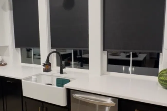 Modern kitchen with black cabinets, white countertops, farmhouse sink, stainless steel dishwasher, and black window shades.