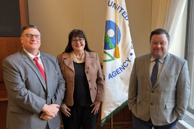 Three professionals posing indoors near a United States EPA flag, dressed in business attire, smiling at camera.
