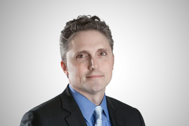 Professional middle-aged man with gray hair wearing a dark suit, blue shirt, and patterned tie on black background