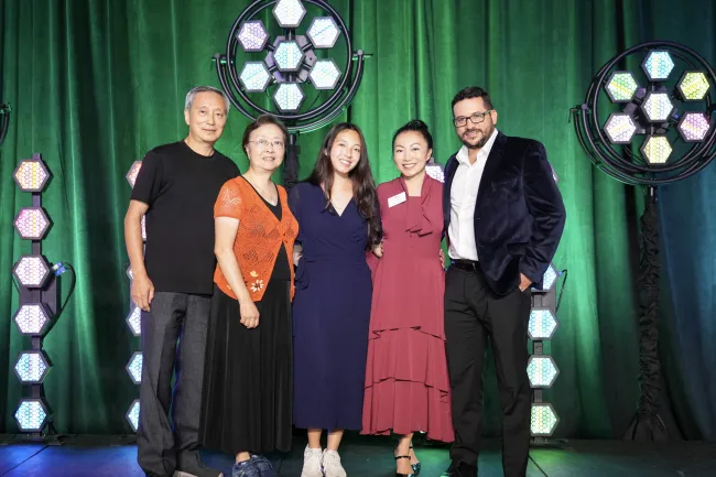 Five people standing together on stage with green backdrop and hexagonal LED lights, smiling at the camera