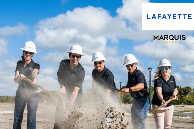 Five people wearing hard hats and black shirts perform a groundbreaking ceremony with shovels on a sunny day.