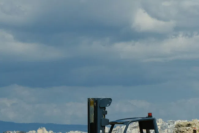 Orange Jungheinrich forklift parked outdoors near stone blocks with city skyline and cloudy sky in background