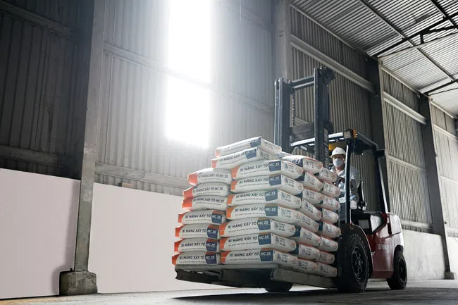 Forklift carrying stacked cement bags inside a warehouse with high windows and industrial walls.