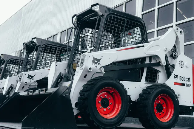 Row of white Bobcat S530 skid-steer loaders with red wheels parked outside a building under overcast sky