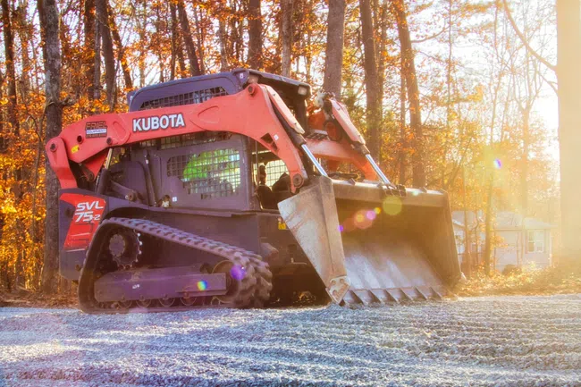 Kubota SVL75-2 compact track loader working on gravel construction site in autumn forest sunset.