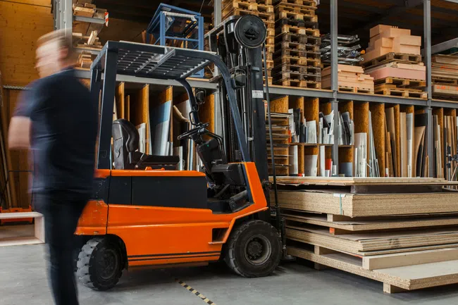 Orange forklift in wood warehouse with stacked plywood and pallets, blurred worker walking by.