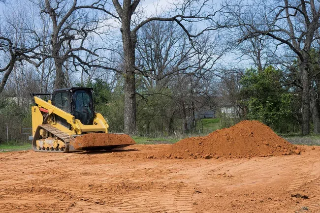 Yellow bulldozer leveling dirt on a cleared land with trees and blue sky in the background
