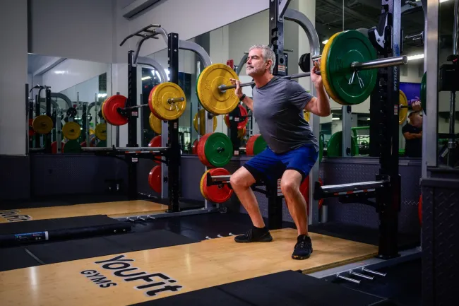 Mature man doing weighted squats with barbell and colorful weights in a modern gym with mirrors.