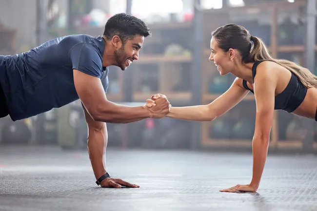 Man and woman doing plank exercise while shaking hands, smiling and working out together indoors.