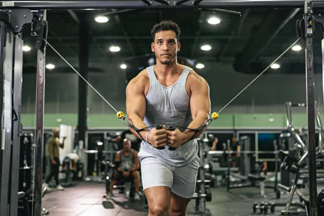 Muscular man in gray tank top and shorts performing cable chest fly exercise in gym
