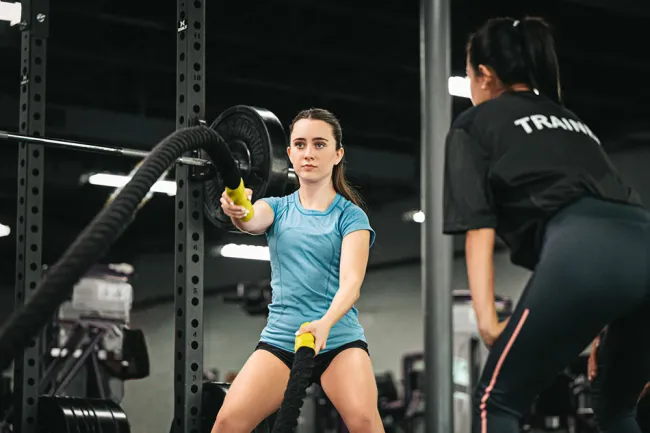 Woman in blue shirt exercises with battle ropes under guidance of female trainer in gym setting