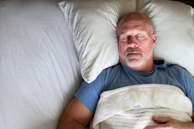 Bald middle-aged man with beard sleeping peacefully on white pillow and blanket wearing a blue shirt.