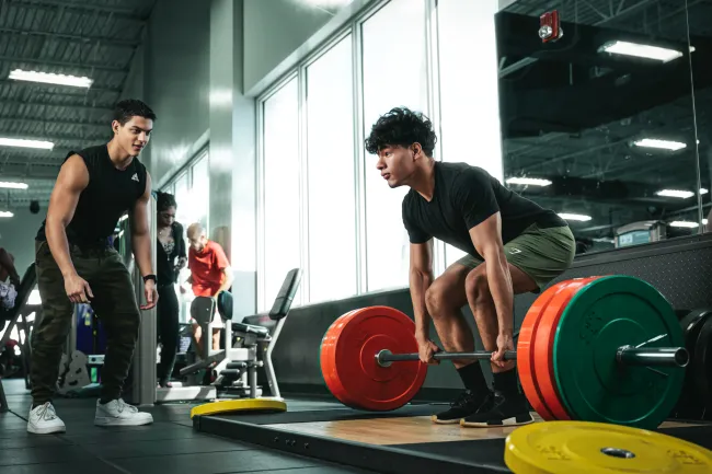 Young man deadlifting heavy barbell at gym while trainer watches and motivates him during workout session.