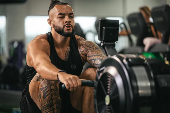 Muscular tattooed man using rowing machine in gym focused on intense workout session.