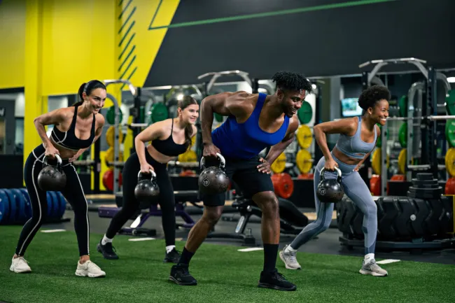 Group of four diverse people exercising with kettlebells in a modern gym with bright yellow walls and equipment.