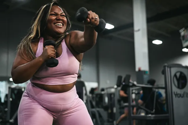 Smiling woman in pink workout clothes lifting dumbbells in a gym during strength training session.