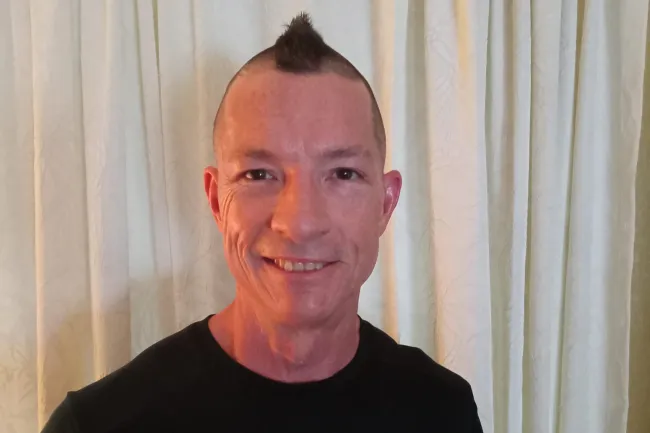 Smiling man with a mohawk haircut wearing a black YouFit Gyms t-shirt in front of white curtains.