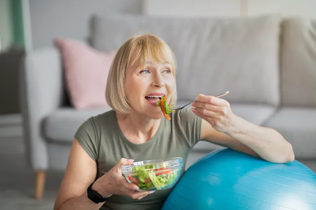 Middle-aged woman eating a fresh salad while sitting next to a blue exercise ball at home.