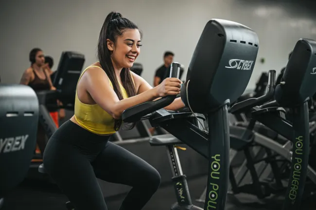 Young woman in yellow sportswear smiling while using a Cybex exercise bike in the gym.