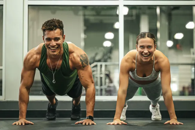 Young man and woman doing push-ups together in a gym with smiles, wearing athletic clothing.
