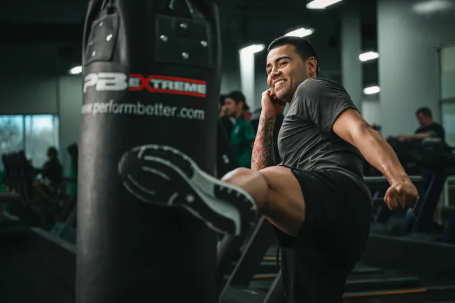 Man in athletic wear kicking a punching bag in a gym with a focused and smiling expression.