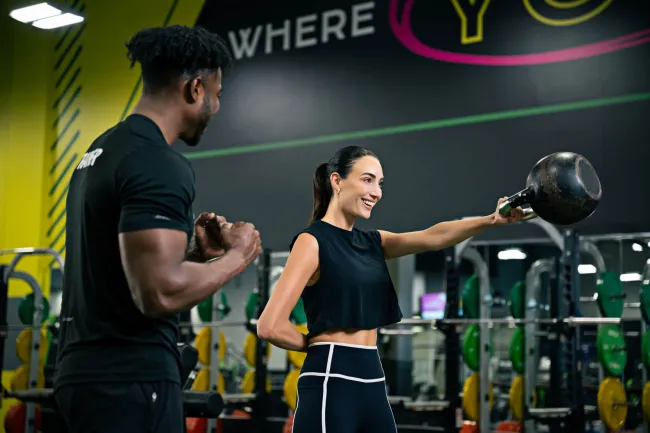 Female lifting kettlebell with male trainer coaching her in gym with weight racks and colorful plates in background