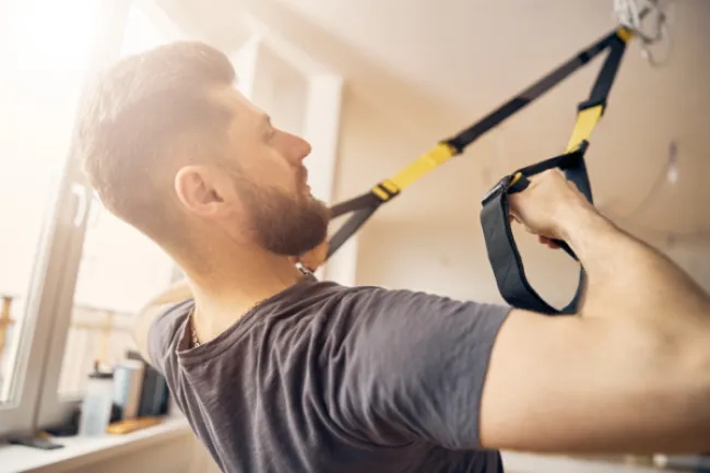 Man with beard exercising using suspension trainer straps in a bright indoor gym space.