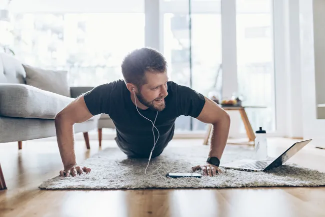 Man doing push-ups at home with earphones, laptop, and water bottle on wooden floor in bright living room.